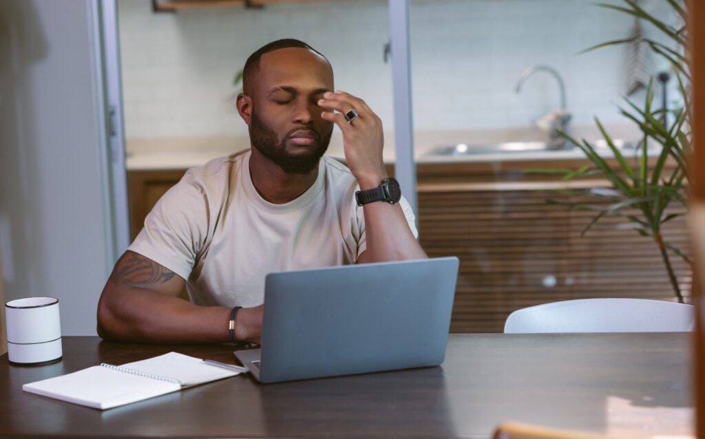 Frustrated African American businessman sit at desk keeping eyes closed, working with laptop at home