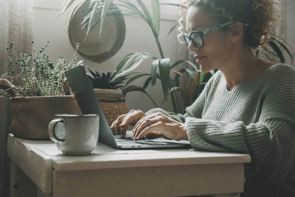 One adult woman writing on laptop at home in green indoor garden workplace.