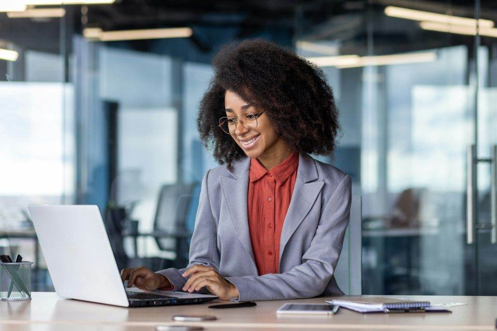 Smiling black woman in eyeglasses using pc while working by equipped table during daytime