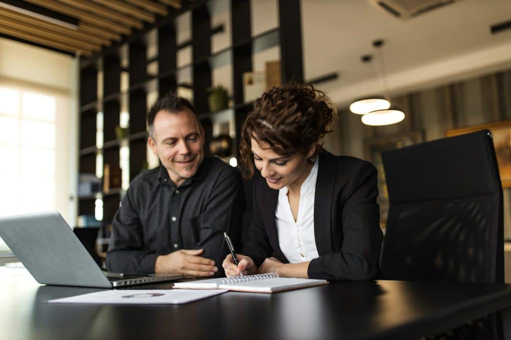 Portrait of a business woman writing in note pad and businessman looking while sitting with laptop.