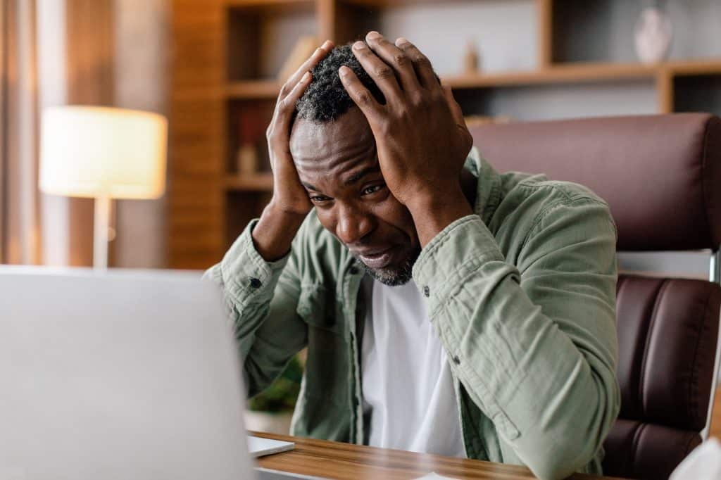 Sad frustrated adult african american guy in casual hold his head with hand watch at laptop, suffer